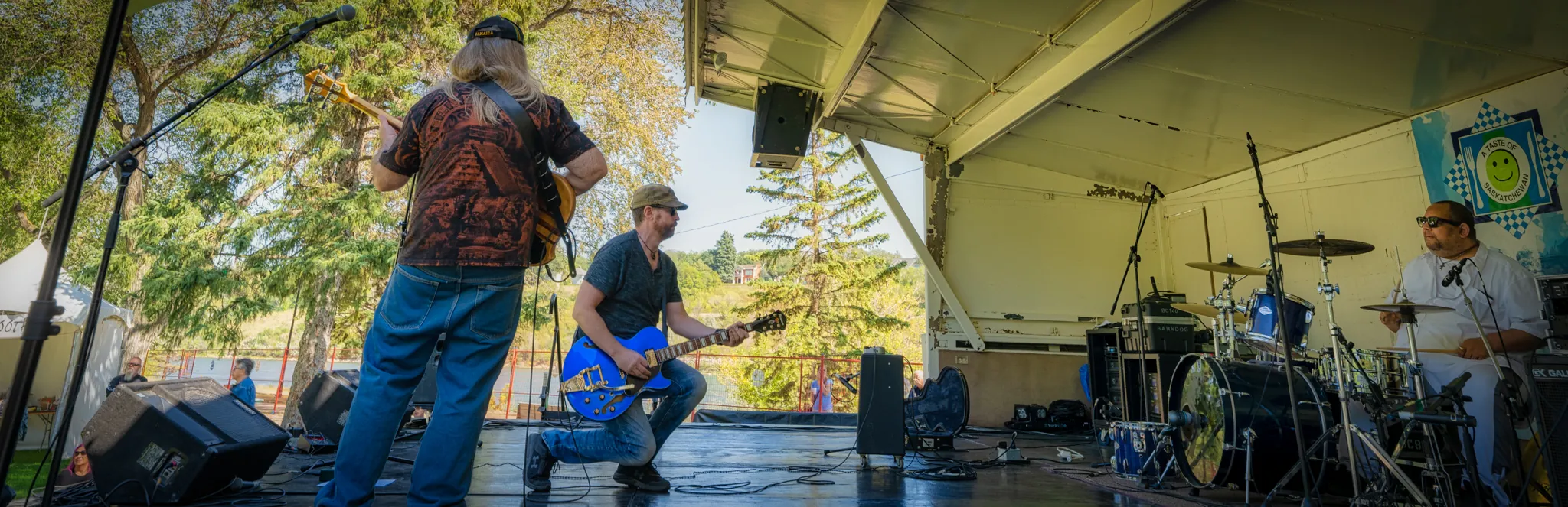 Michael Lander kneeling with a blue electric guitar on an outdoor stage beside the river